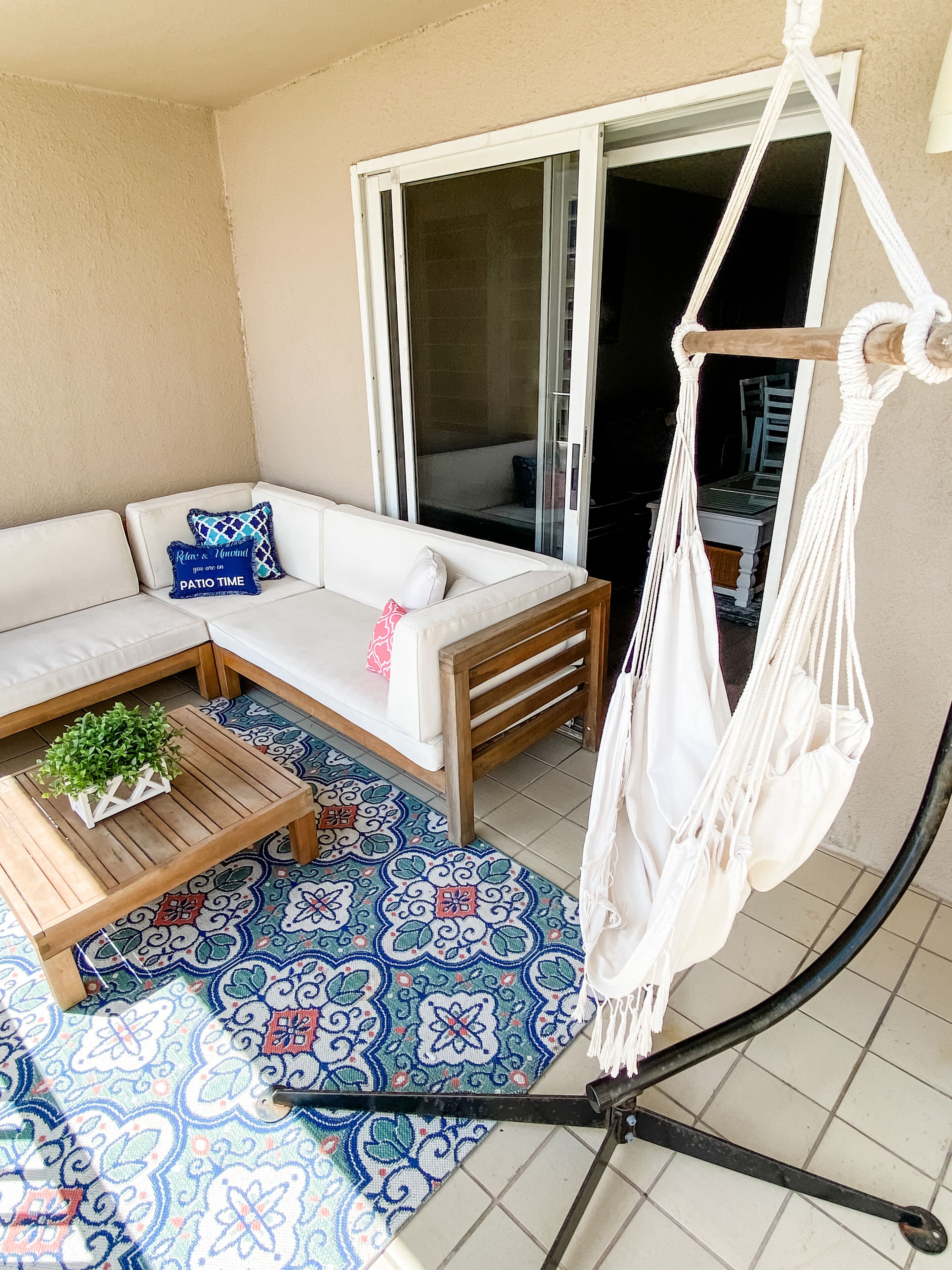 A balcony with a white sofa, a wooden coffee table, a hanging macrame chair, and a patterned floor mat.