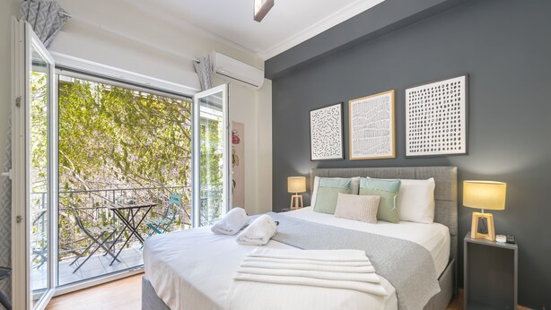 A bedroom with a large bed, two bedside tables with lamps, a balcony with a table and chairs, and artwork on the wall. - Elegant Suite by CloudKeys (Athens)