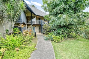 Set amidst lush greenery, this two-story building features a balcony on the upper floor, accessible via a concrete pathway.