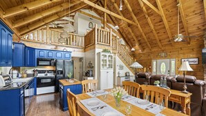 Una cocina amplia con armarios azules, una zona de comedor con mesa de madera dispuesta para cuatro personas y una sala de estar acogedora con un sofá de cuero marrón.