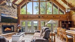 Interior acogedor de una cabaña con chimenea de piedra, sofá de cuero, mesa de comedor de madera y ventanas amplias con vistas al bosque.