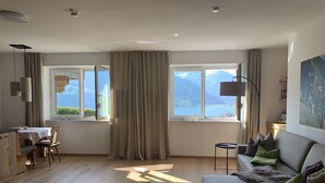 Ein großzügiger Wohnraum mit Holzboden, einer Couch, einem Esstisch mit Stühlen und großen Fenstern mit Blick auf Berge und einen See.