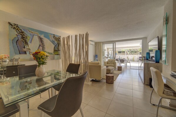 A modern living room with a glass dining table, comfortable seating, a large painting, and a view through a glass door. - Luxury Apartment, High-Speed Wi-Fi, Sunny Terrace, Heated Pool, Marazul (Adeje)