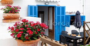 A terrace with potted red flowers, a table with a plate and a glass, and blue shutters on the building.