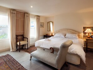 A neatly arranged bedroom with a large bed, a chair, a bedside table with a lamp, and a bookshelf.