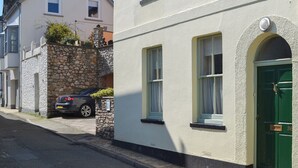 Straßenansicht mit einer Reihe weißer Gebäude, eines mit einer grünen Tür und ein weiteres mit einer Steinwand und einem parkenden Auto.