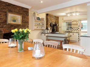A dining area with a wooden table, chairs, and a vase with yellow flowers. There is a kitchen in the background with various appliances and utensils.