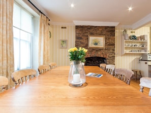 Een eetkamer met een lange houten tafel, stoelen, een vaas met bloemen, een open haard en een boekenkast.