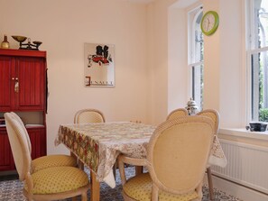 Een zithoek met een tafel bedekt met een kantkleed, omringd door rieten stoelen met gele kussens. Een rode kast met messing decoratie staat links, en een groene klok hangt rechts aan de muur.