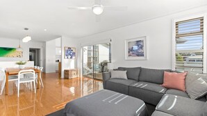 Un salon moderne avec un canapé gris, une table à manger avec des chaises, un ventilateur de plafond et une grande fenêtre avec des stores.