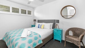 Une chambre à coucher avec un lit, un ventilateur de plafond, un miroir rond, une chaise en osier et une table de chevet de couleur turquoise.
