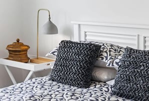 Una cama con ropa de cama estampada, una lámpara de noche y un objeto decorativo de madera sobre una mesita blanca.
