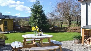 A wooden picnic table with benches, a vase with yellow flowers, and a bottle of wine on a gravel surface. - North Lodge Gate House (Muir of Ord)