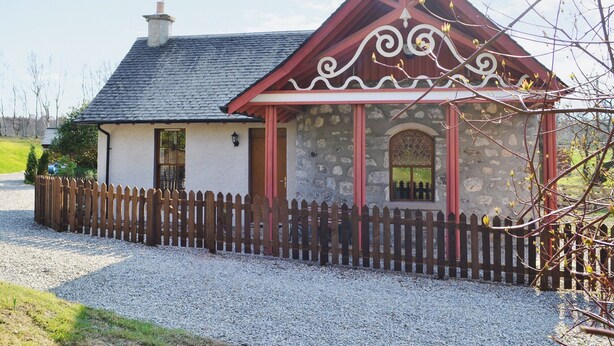 A stone building with a red roof and a wooden picket fence. - North Lodge Gate House (Muir of Ord)