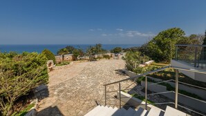 Un chemin de pierre mène à un escalier doté d’une rampe en métal, le tout étant entouré de verdure et offrant une vue sur la côte.