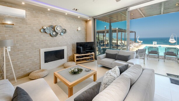 Un salon moderne doté d’une cheminée, d’un téléviseur à écran plat et de sièges confortables. Une grande porte vitrée donne accès à un balcon avec piscine et vue sur l’océan.