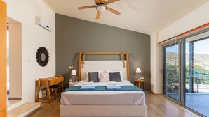 Une chambre moderne avec un grand lit, une tête de lit en bois, des tables de chevet avec des lampes, un ventilateur de plafond et une vue sur une colline grâce à une porte-fenêtre coulissante.