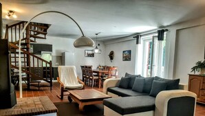 Un vaste salon comprenant un canapé, un fauteuil et une table à manger. Un escalier avec des rampes en bois mène à l’étage supérieur.
