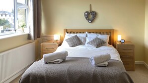 Un lit bien fait, avec deux serviettes enroulées, une décoration murale en forme de cœur et une horloge de chevet posée sur une table de nuit en bois.