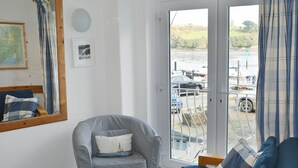 A room with a grey armchair, a blue sofa, a wooden floor, a map on the wall, and a view of a dock with boats and cars.