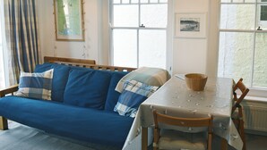 A room with a blue sofa, a dining table with a bird-patterned tablecloth, and a window with a blue and white valance. - 1 bedroom accommodation in Fore St (Fore St)