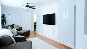 A modern living room with a grey sectional sofa, a ceiling fan, a flat-screen TV mounted on the wall, and a wooden floor.