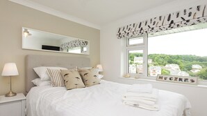 Uma cama bem arrumada com lençóis brancos, um espelho grande acima e uma janela com vista para casas e vegetação.