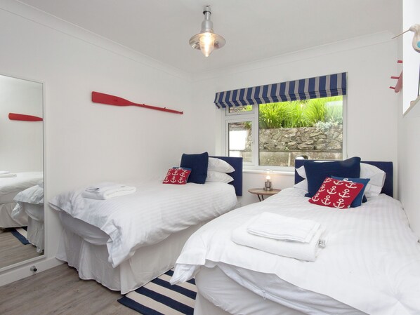 Habitación de hotel con dos camas, ropa de cama blanca, almohadas rojas y azules, una alfombra a rayas, una mesita con lámpara, una ventana con vistas a zonas verdes y una pala roja en la pared.