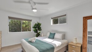 Quarto com cama, ventoinha no teto, duas mesas de cabeceira, um criado-mudo, uma planta em vaso e duas janelas.