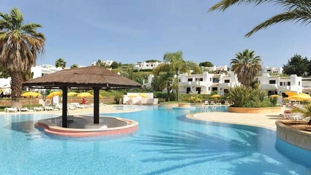 Ein Swimmingpool mit einem Reetdach-Pavillon, Liegestühlen und Sonnenschirmen.