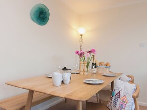 A wooden dining table with white plates, cups, and a bottle of champagne. There are pink flowers in a vase, a basket of bread, and a lamp on the table.