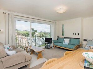 Un salón con sofá, mesa de madera y televisor. Posee un balcón con vistas a casas y follaje.