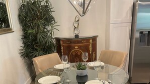A dining area with a round glass table, four upholstered chairs, a decorative chandelier, a potted plant, and a white cabinet.
