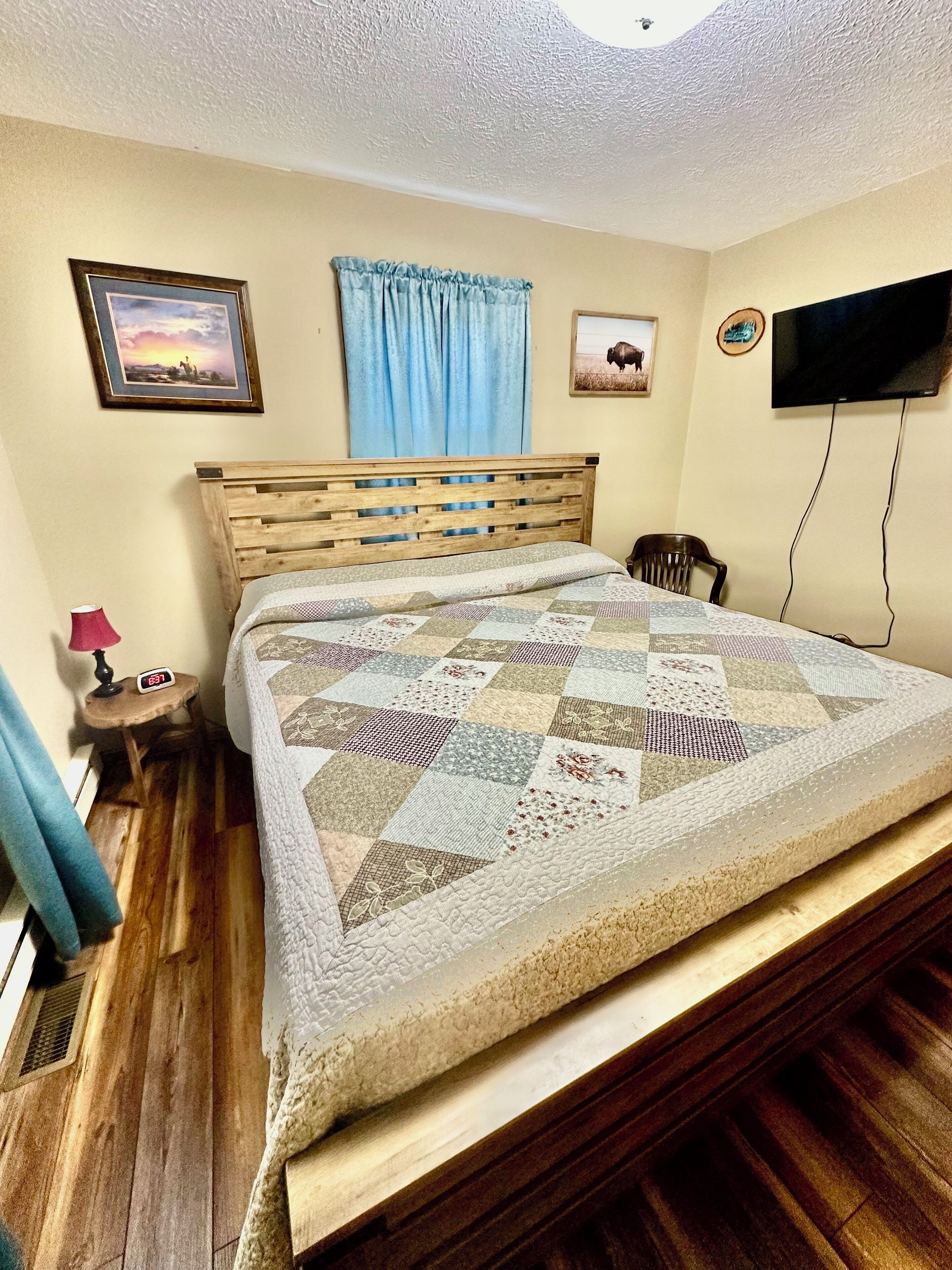 Une chambre à coucher avec un lit en bois, une courtepointe, une petite table de chevet avec une lampe, un cadre photo, un téléviseur et une fenêtre avec des rideaux.