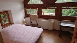 A bedroom with a bed, a desk, a chair, and a radiator. There are windows providing a view of greenery outside.