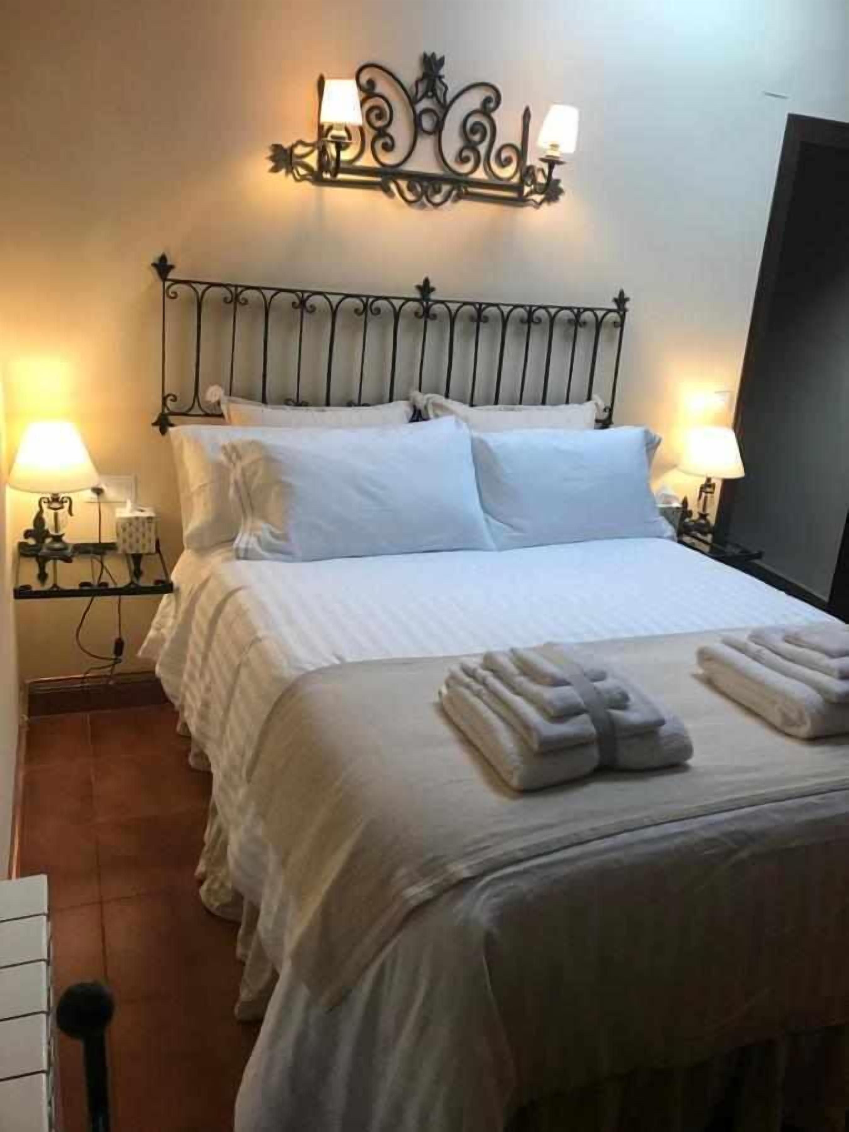 A neatly made bed with white linens and towels, a decorative wall-mounted light fixture, and a simple bedside table with a lamp.