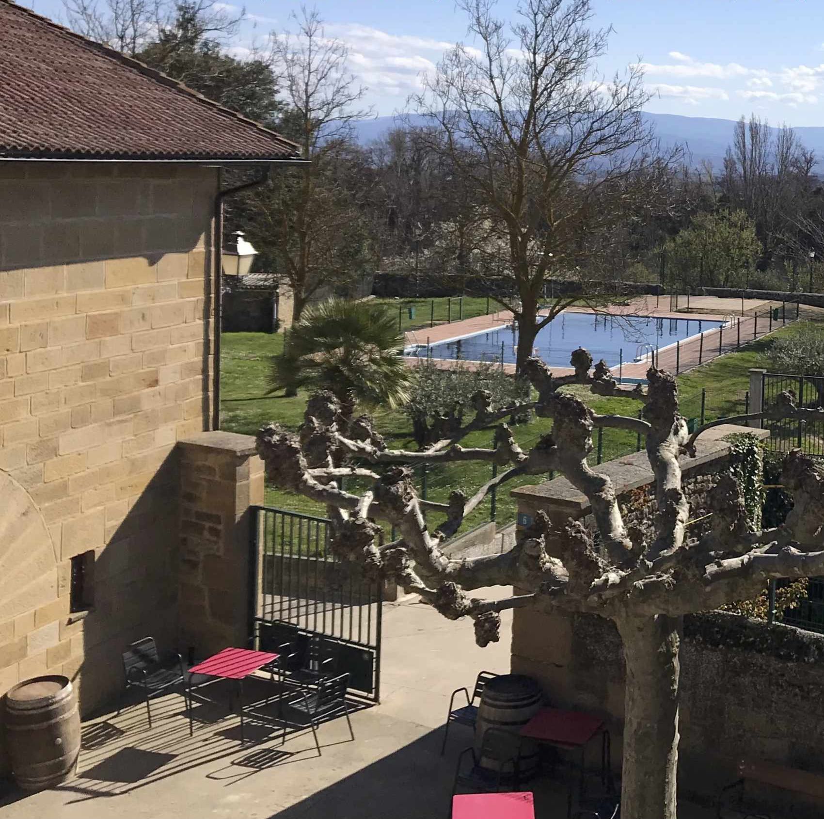 A view from a balcony overlooking a swimming pool, with a stone building and patio furniture visible.