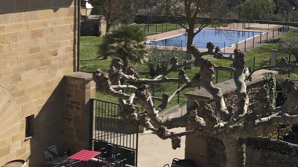 A view from a balcony overlooking a swimming pool, with a stone building and patio furniture visible.