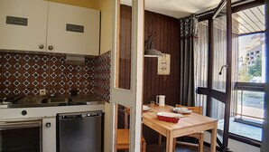 A compact kitchen with a stove, sink, and refrigerator. There is a dining table with chairs and a window with curtains. - travelski home classic - Résidence Origanes - Studio 2 personnes (Les Menuires)