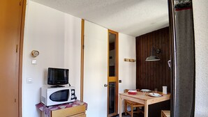 A compact room with a wooden dresser, a microwave, a small table with a chair, and a door leading to another room. - travelski home classic - Résidence Origanes - Studio 2 personnes (Les Menuires)