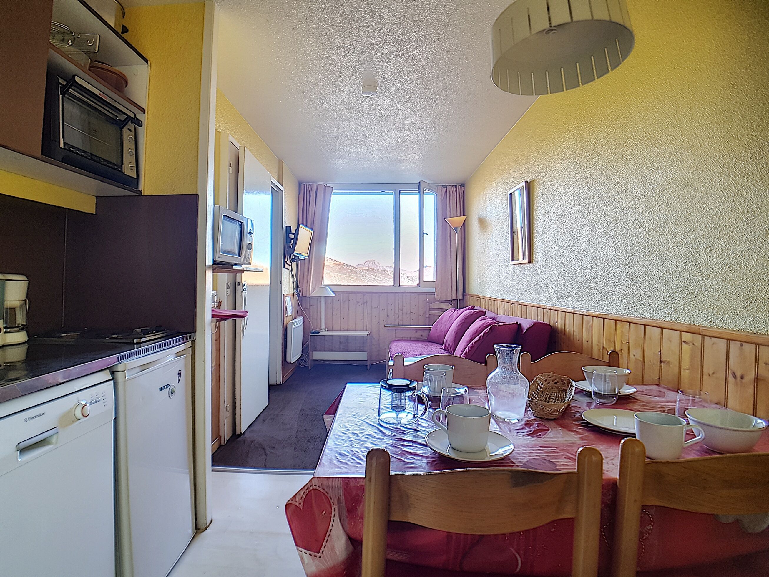 Ett kompakt kök med mikrovågsugn, brödrost och kaffebryggare. Ett matområde med ett bord för två personer, en soffa och utsikt över omgivningarna.