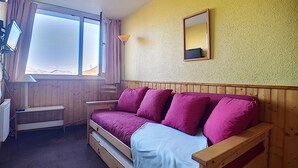 Ein Zimmer mit einem hölzernen Bett, einem kleinen Schreibtisch, einem Stuhl, einem Fenster mit Vorhängen und einem Spiegel an der Wand.