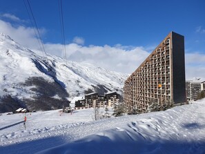 Un hotel moderno con una fachada geométrica distintiva, ubicado en una zona montañosa nevada con pistas de esquí al fondo.