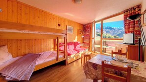 A room with wooden walls, bunk beds, a dining table with chairs, and a view of mountains through a large window. - travelski home classic - Résidence Grande Masse - Studio 3 personnes 709 (Les Menuires)