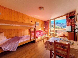 A room with wooden walls, bunk beds, a dining table with chairs, and a view of mountains through a large window. - travelski home classic - Résidence Grande Masse - Studio 3 personnes 709 (Les Menuires)