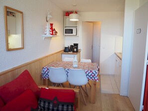 A compact living space with a kitchenette, a dining area with two chairs, a sofa with red cushions, and a microwave above the counter.