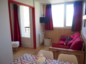 A cozy room with a red sofa, a small white chair, a table with a patterned tablecloth, and a TV mounted on the wall.