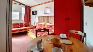 Ein Zimmer mit einem Esstisch, einer Couch mit gemusterten Kissen, einer roten Tür und einem an der Wand befestigten Fernseher.