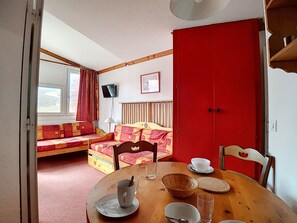 Ein Zimmer mit einem Esstisch, einer Couch mit gemusterten Kissen, einer roten Tür und einem an der Wand befestigten Fernseher.