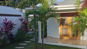 A modern house with a wooden door, a small porch, and a well-maintained garden with palm trees and various plants. - 5 bedroom villa, large swimming pool 250m from the sea (Las Terrenas)
